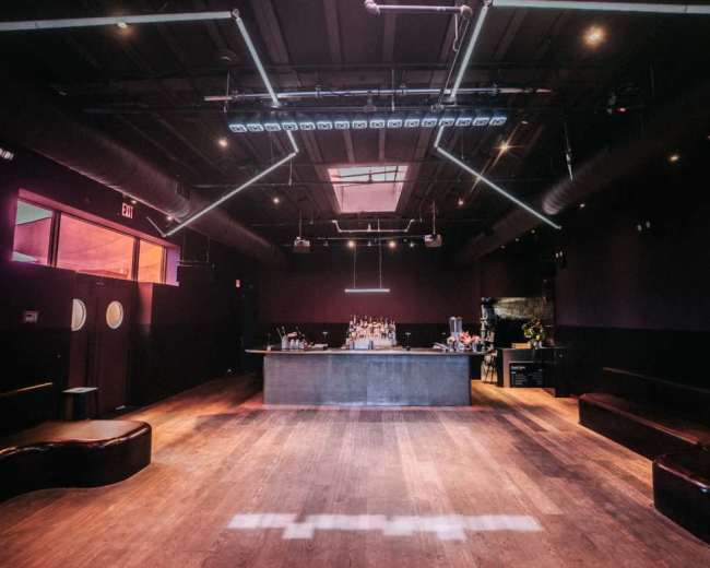 The image shows a dimly lit bar area with a wooden floor, sleek bar counter, and seating along the walls.
