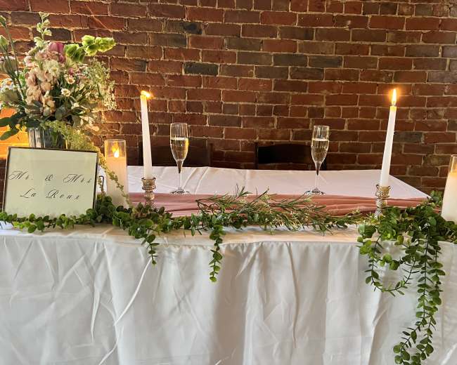 A decorated table featuring floral arrangements, candles, and champagne glasses is set against a brick wall.