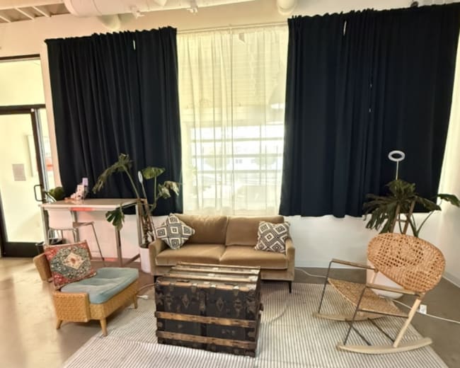The image shows a cozy lounge area featuring a brown sofa, a wooden coffee table, and various plants, with large windows covered by dark curtains.