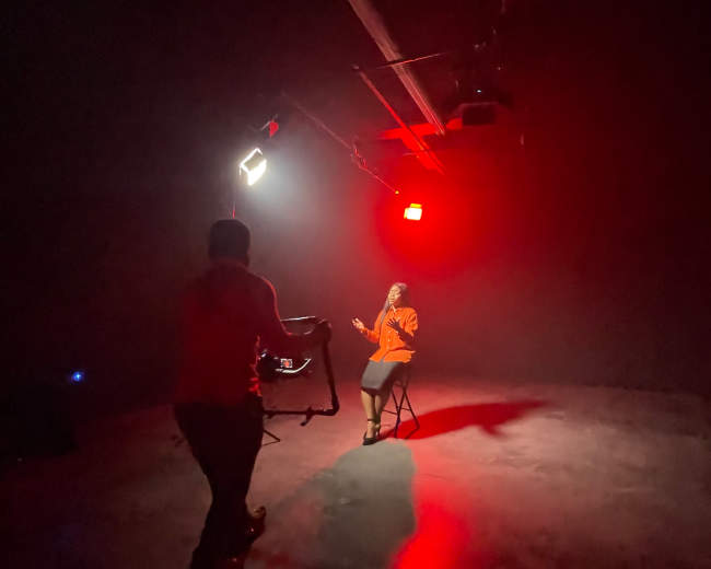 A person sits on a chair in a dimly lit studio, illuminated by red lights, while another person operates a camera nearby.