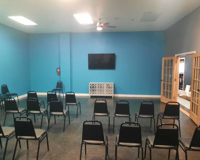 A brightly colored room contains rows of black chairs facing a mounted television on a turquoise wall.