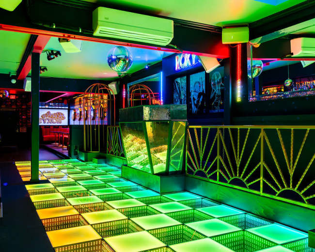 The image shows a brightly lit nightclub interior featuring a colorful illuminated dance floor with a mirrored disco ball and stylish decor.