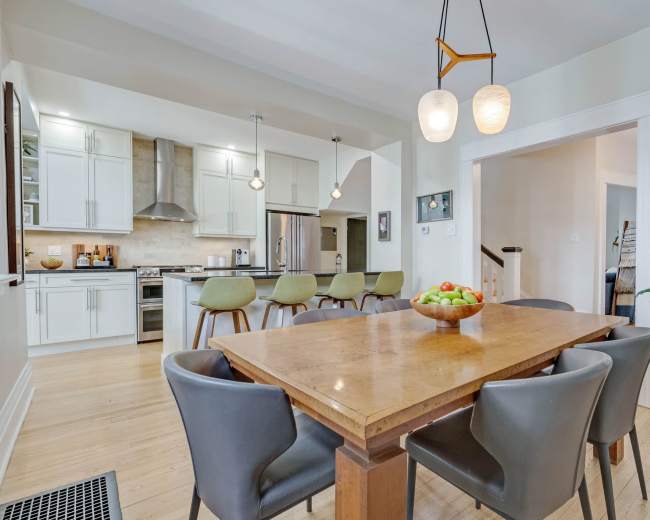 The image shows a modern kitchen and dining area featuring a wooden table with gray chairs, pendant lighting, and stainless steel appliances.