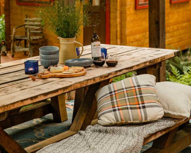 A rustic wooden outdoor dining table is set with food and drinks, nestled beside a cozy cabin in a forested area.