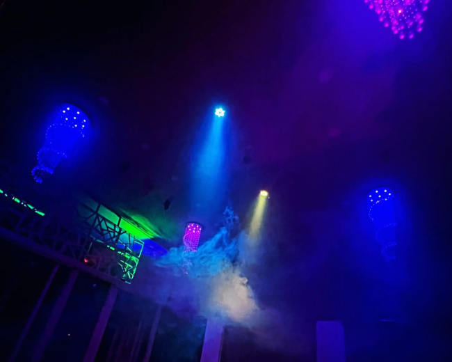 A dimly lit interior features colorful lights and smoke, with chandeliers hanging from the ceiling.