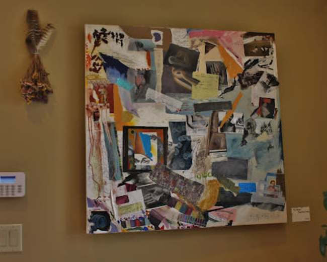 A collage artwork composed of various mixed media elements is displayed on a wall next to a light switch and a security panel.