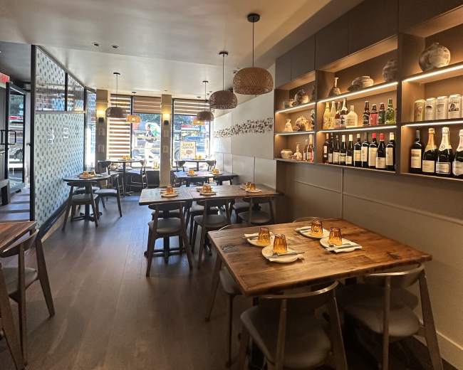 The image shows a modern restaurant interior with wooden tables, hanging light fixtures, and a shelf displaying bottles and decorative items against a neutral wall.