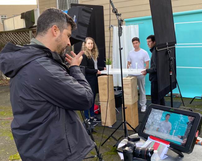 A photographer sets up a photoshoot with three models in front of a turquoise backdrop, surrounded by lighting equipment and a monitor displaying the shot.