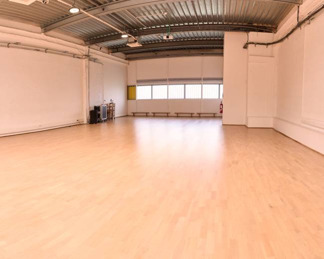 The image shows a spacious, empty room with wooden flooring and large windows providing natural light.