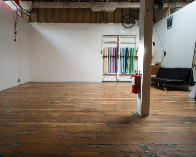 An empty room features wooden floors, a white painted wall, a couch, a fire extinguisher, and colorful materials hanging on another wall.
