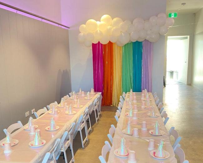 A long, rectangular table is set with pastel-colored party hats and plates beneath a rainbow curtain and decorative balloon cloud.