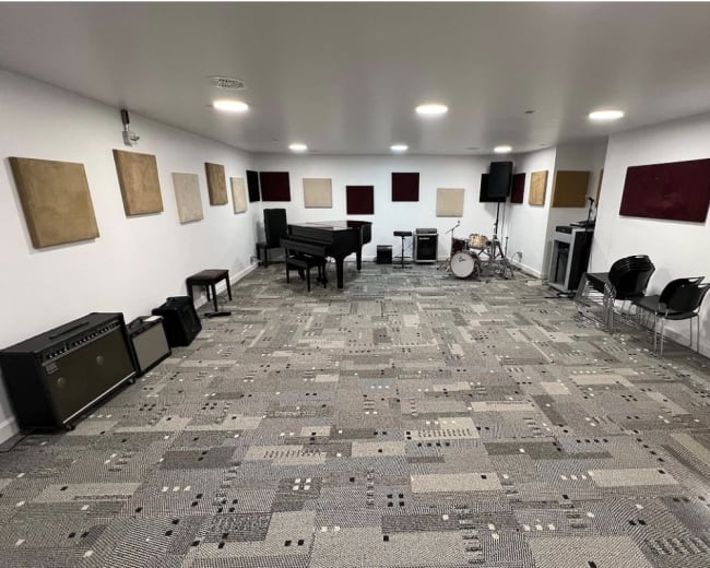 The image depicts a spacious rehearsal room equipped with a grand piano, drum set, amplifier, and several empty chairs arranged against the walls adorned with sound-absorbing panels.