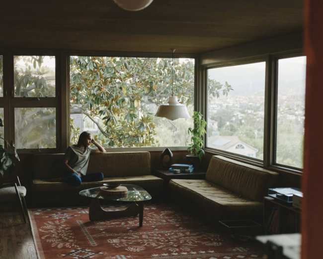 A person sits thoughtfully on a couch in a spacious living room with large windows overlooking a view of greenery and distant hills.