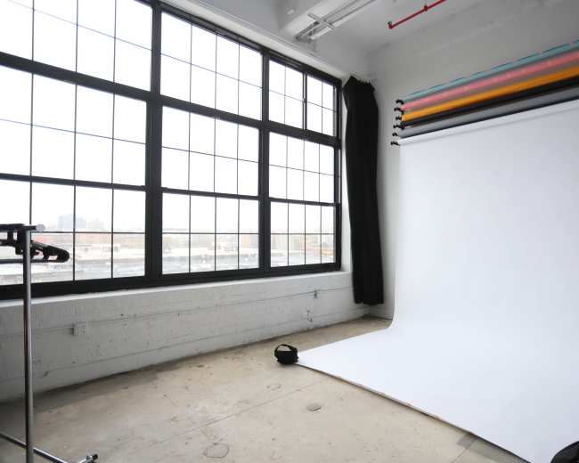 The image depicts a spacious photography studio with large windows, a roll of white backdrop paper, and a clothes rack in the corner.