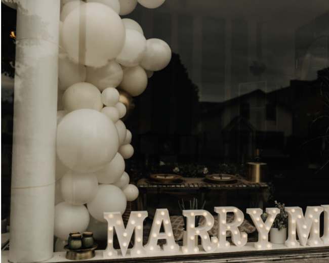 A window display features a "MARRY ME" sign illuminated with lights, accompanied by a cluster of white balloons and decorative elements.