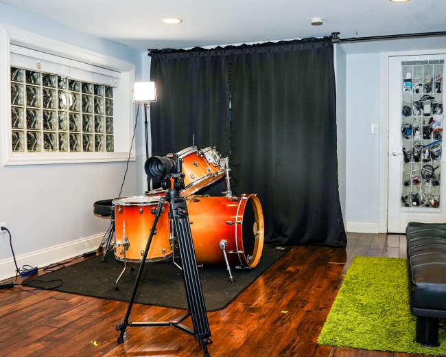 The room features a set of drum equipment on a black rug, a tripod with a camera, two lighting stands, and a black curtain in the background.