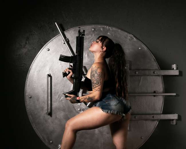 A woman in denim shorts and high heels poses confidently with an assault rifle in front of a large safe door.