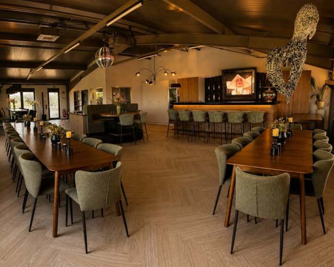 The image shows an interior of a dining space with multiple large tables arranged neatly, surrounded by upholstered chairs and featuring a bar area in the background.