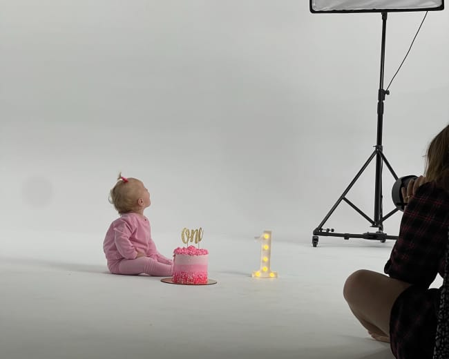 A baby sits on the floor in front of a pink birthday cake with a candle while a softbox light is set up nearby.