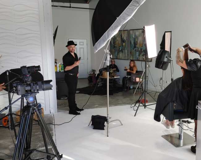 A hairstylist is working on a client's hair in a studio setting, while a photographer and other individuals observe in the background.