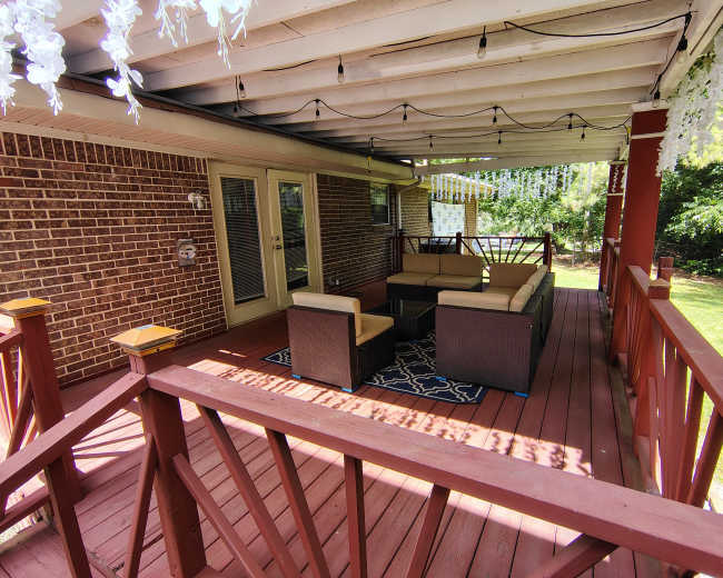 The image shows a covered wooden deck with a seating area consisting of a brown wicker sofa and a table, surrounded by greenery.