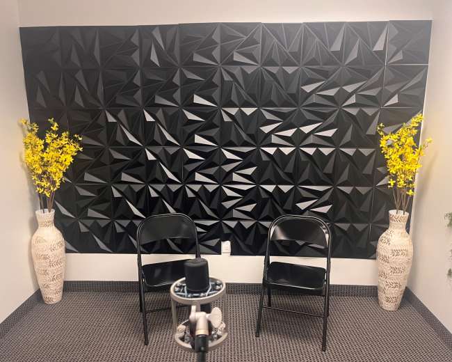 The image shows a small room with a black geometric accent wall, two black folding chairs facing each other, and decorative vases containing yellow flowers on either side.