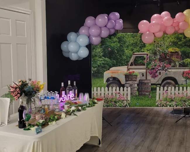 A table with floral arrangements and drinks is set up in front of a backdrop featuring an antique truck surrounded by greenery and a white picket fence, adorned with a colorful balloon arch.