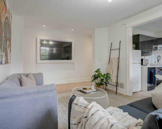 A modern living room features a gray sofa, a round coffee table, and a wall-mounted TV, with an open view toward a kitchen area.