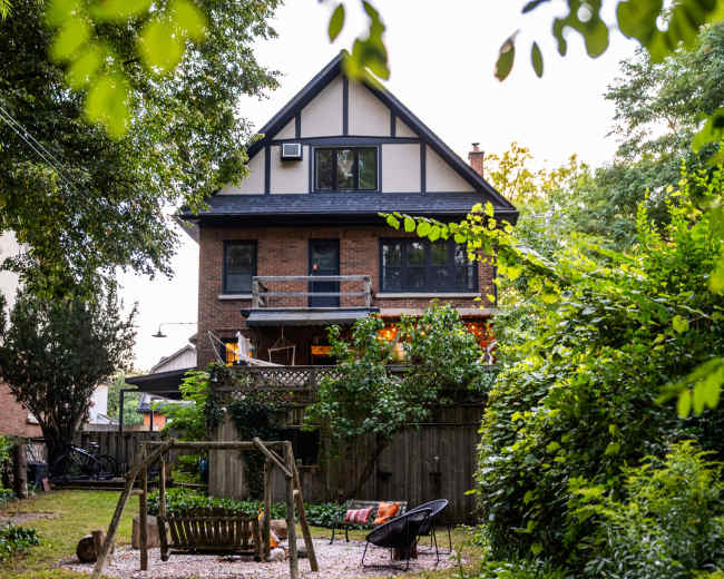 A two-story house with a balcony and a backyard featuring a swing set is surrounded by trees and greenery.