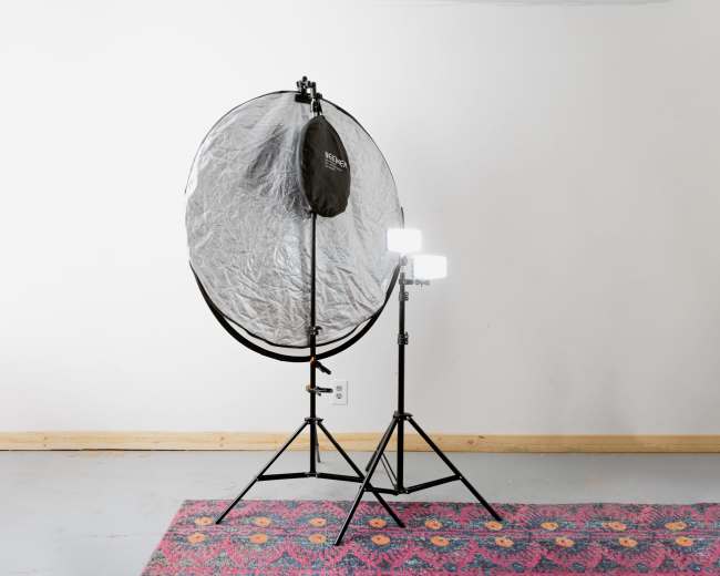 A photography setup featuring a silver reflector on a stand and two LED light panels on tripods, positioned on a patterned rug in a minimalistic space.