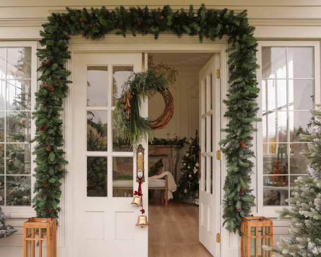 The image shows a decorated entrance with evergreen garlands, a wreath, and two lanterns on either side, leading into a room with Christmas trees visible inside.