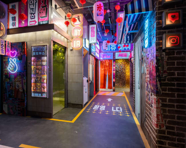The image shows a vibrant commercial alleyway illuminated by neon signs and lanterns, featuring storefronts with various graphics and colorful lighting.