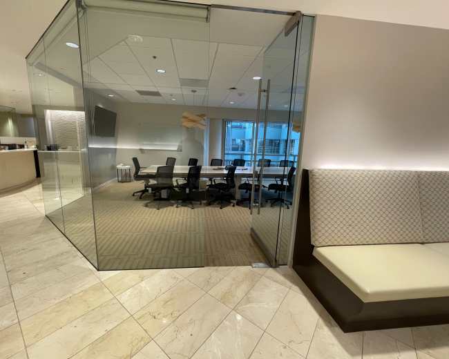 The image shows a modern office space with a glass-walled conference room containing several chairs and a large table, adjacent to a reception area.