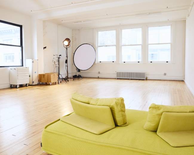 The image shows a bright, empty studio space with wooden floors, large windows, a green couch, and lighting equipment in the corner.