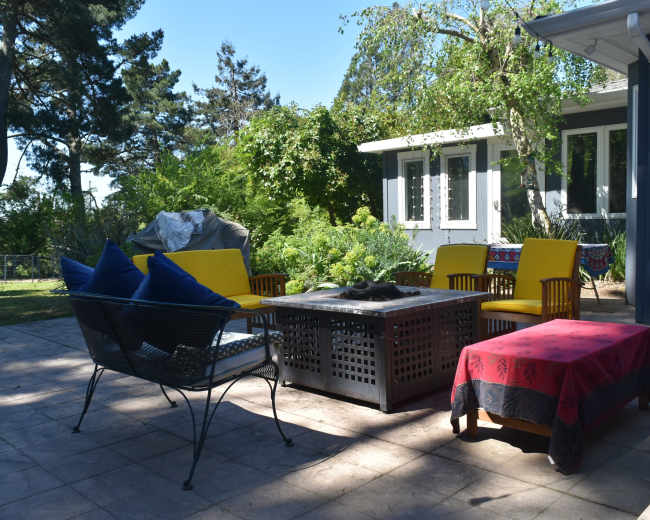 A patio area features a table with a fire pit surrounded by yellow chairs and a red bench, with greenery and a house in the background.
