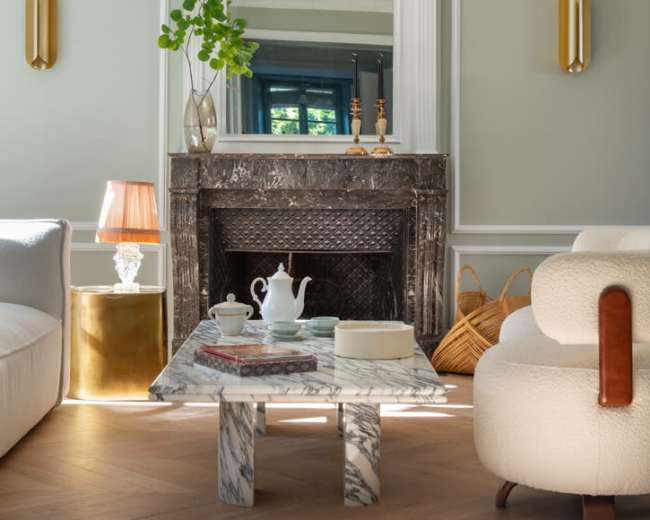 The scene features a modern living room with a marble coffee table, a decorative fireplace, and soft seating arrangements, highlighted by natural light and elegant decor.