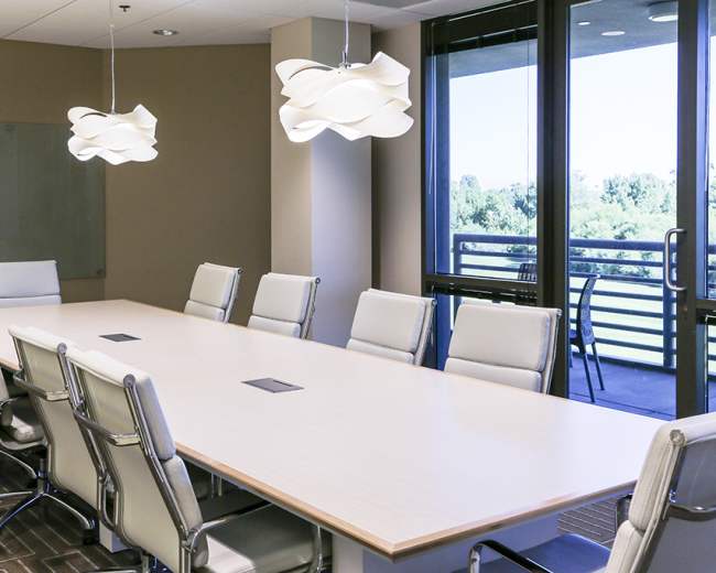 A spacious conference room features a long wooden table surrounded by modern white chairs, with large windows providing views of greenery outside.