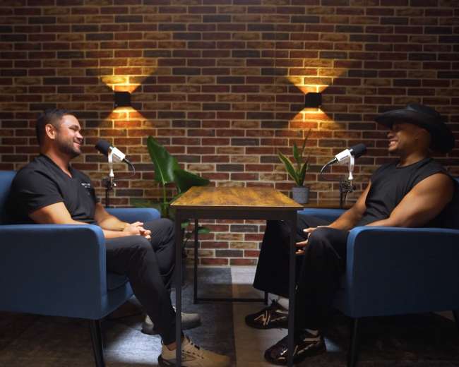 Two men sit in blue chairs across from each other at a wooden table, with brick walls and soft lighting in the background, equipped with microphones for a conversation.