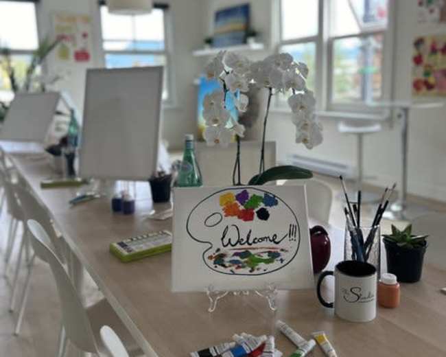 A brightly lit room features a long table set with art supplies, small plants, and a welcoming sign surrounded by chairs and easels.