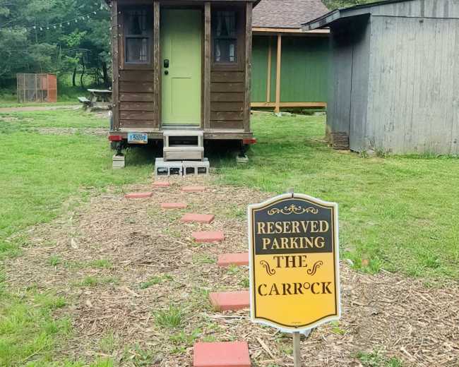 The image shows a small, wooden cabin with a green door set on a trailer, accompanied by a sign that reads "Reserved Parking The Carrock," amidst a grassy area surrounded by trees.