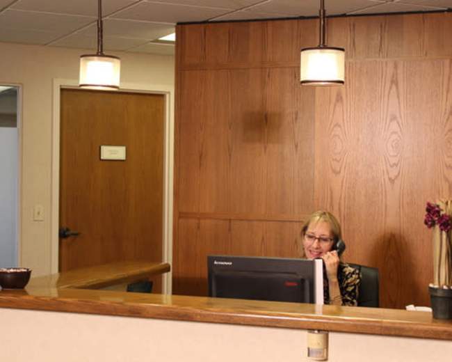 A receptionist sits at a desk in an office, speaking on the phone while using a computer monitor.
