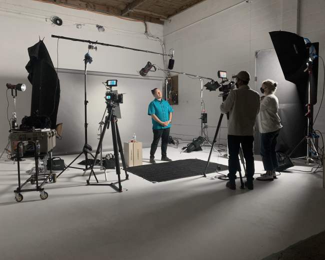A video shoot is underway in a studio featuring multiple cameras, lighting equipment, and a person standing in front of a backdrop with crew members observing.