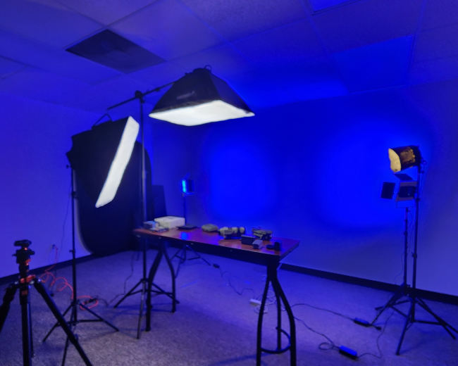 The image shows a dimly lit studio setup with blue lighting, featuring a table and various equipment, including light stands and a backdrop.