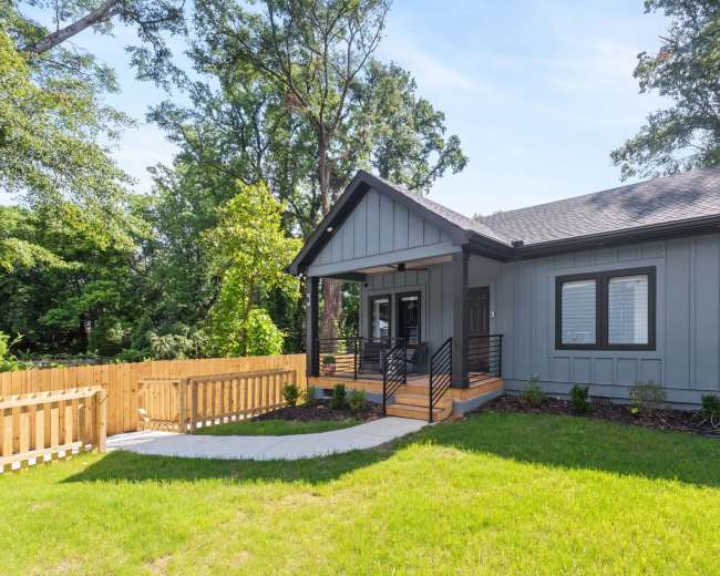 The image shows a contemporary single-story house with a gray exterior, a wood front porch, and a landscaped yard surrounded by a wooden fence.