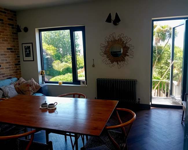 A dining area features a wooden table surrounded by chairs, large windows overlooking a garden, and a doorway leading to a balcony.