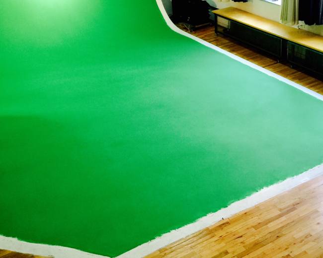 A large green backdrop is set up in a room with wooden floors and windows, while a person stands nearby.