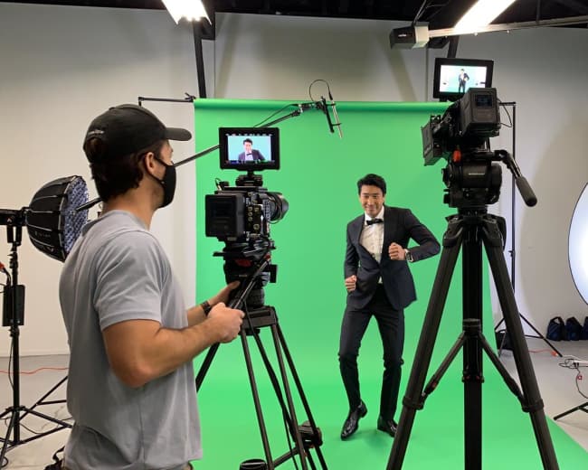 A man in a suit dances in front of a green screen while a camera operator captures the scene with multiple cameras and lighting equipment arranged around them.