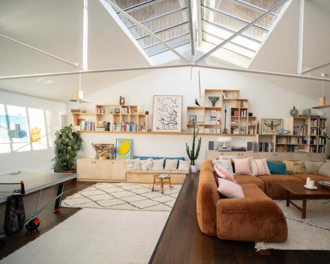 The image shows a spacious, well-lit living room featuring a large sofa, bookshelves, indoor plants, and a table tennis setup.