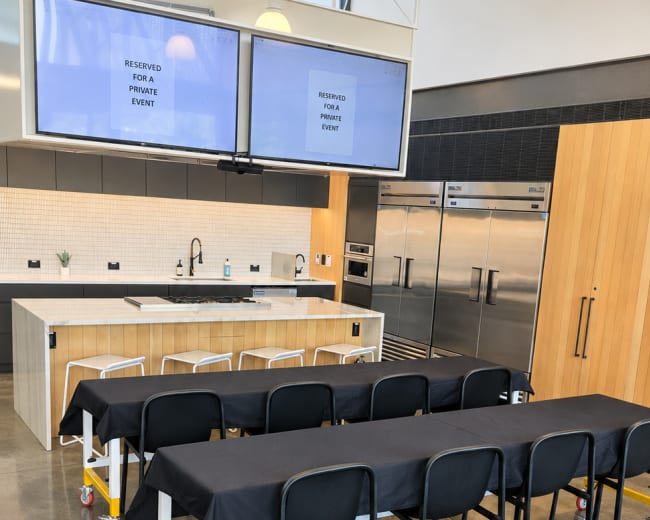 The image shows a modern event space with rows of black-covered tables, a kitchen area in the background, and two large screens displaying "RESERVED FOR PRIVATE EVENT."