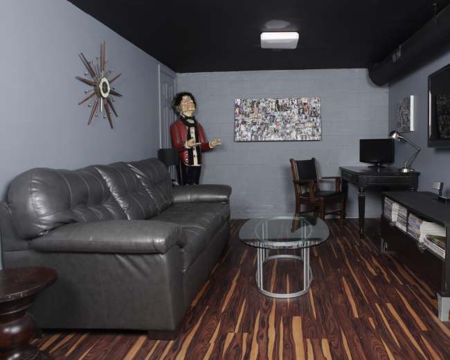 The room features a gray wall, a black leather couch, a glass coffee table, a wooden chair, a desk with a computer, and a life-sized mannequin dressed in a suit.